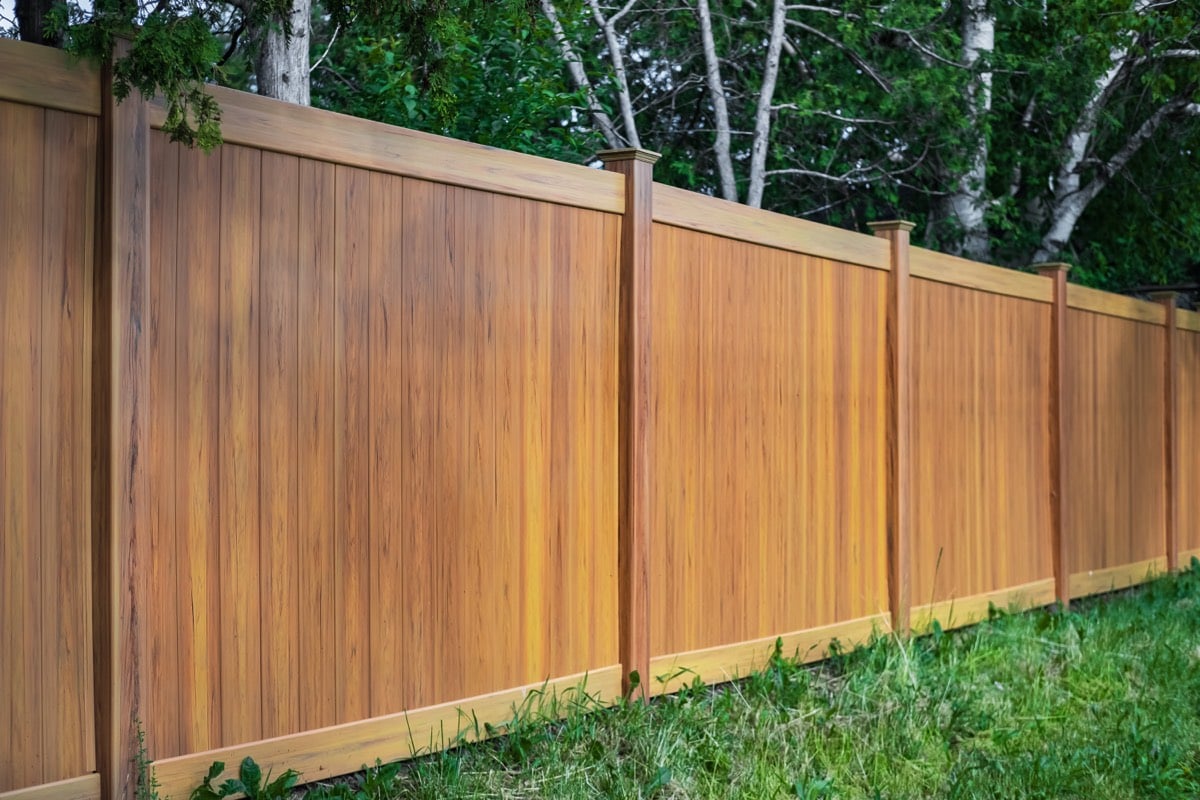 A cedar-tone privacy fence with flat-top posts along a grassy backyard edged by trees